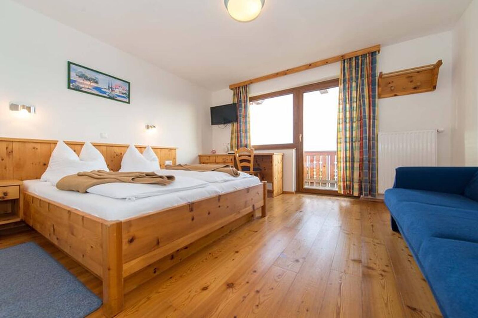 Cozy hotel room featuring wooden decor, a double bed, desk, TV, and colorful curtains, with a balcony view.