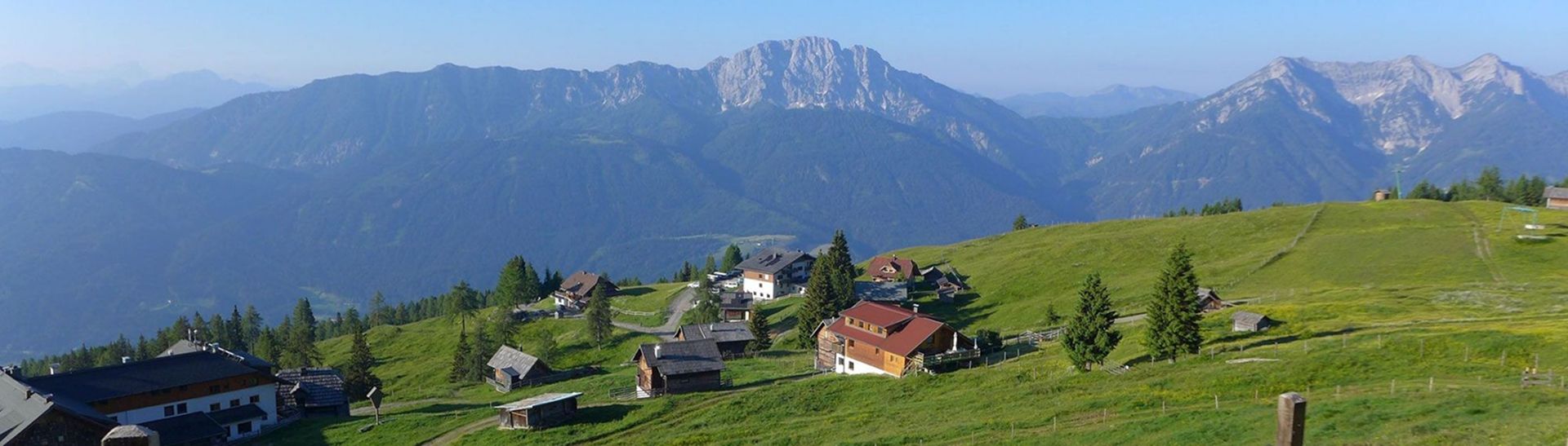 A panoramic view of lush green hills dotted with rustic homes, framed by majestic mountains under a clear blue sky.