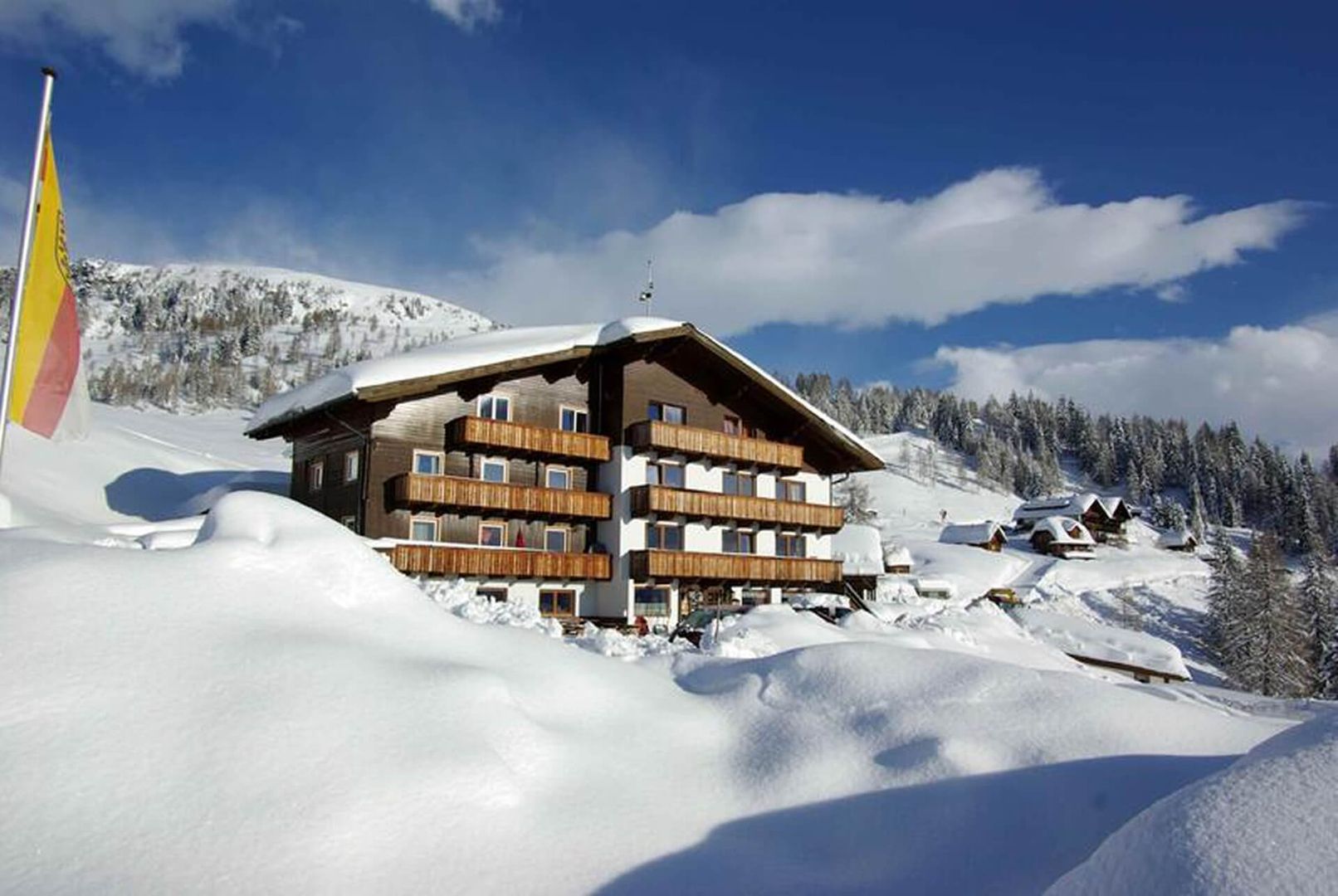A snowy mountain landscape featuring a wooden lodge surrounded by deep snow, with distant peaks and a clear blue sky.